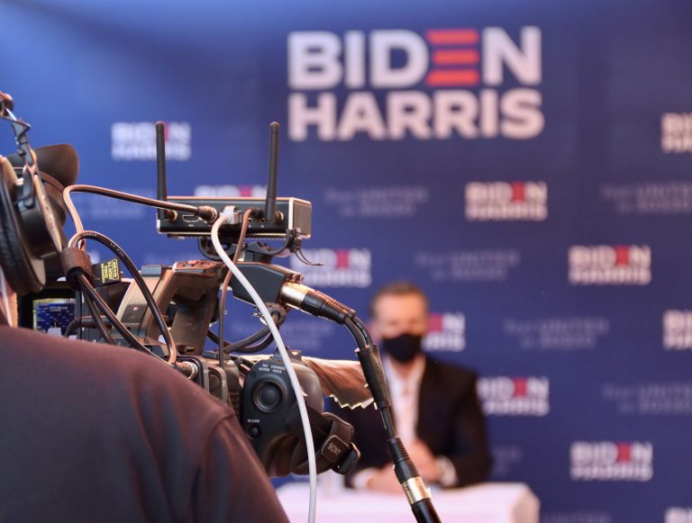 Camera filming a masked speaker at a Biden-Harris event
