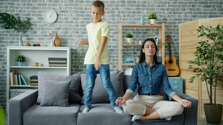 Boy standing on a couch while a woman sits cross-legged meditating beside him in a living room