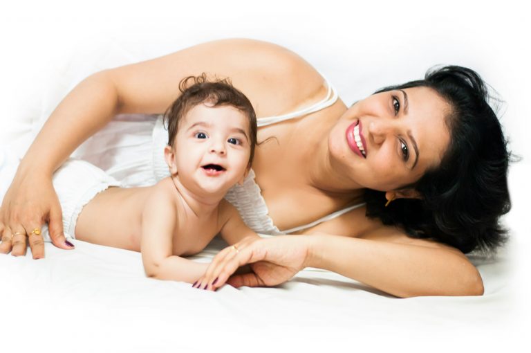 Mother lying with her baby on a white bed, both smiling at the camera